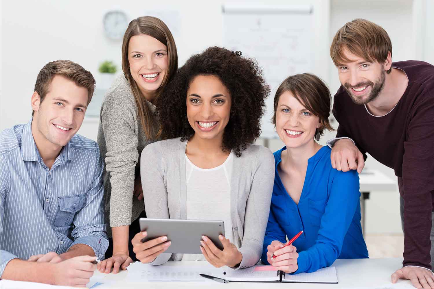A group of five people smile while gathered around a table during a meeting. One person in the center is holding a tablet.